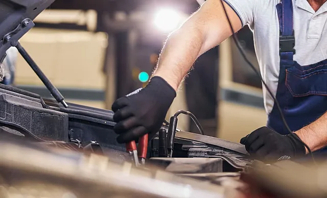 Autoreparatur – Arme eines Mechanikers in blauer Latzhose und schwarzer Handschuhe, der mit Klemmen an einem Motorraum arbeitet.